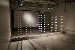 Two of the works in this series, Wall, and Gate seek to locate the body in a place of yearning anticipation, of wanting to reach for the other side, beyond the barrier and border. In the first of these, Wall, the room for the installation is divided into two halves. The viewer can enter into one side or the other, but once there cannot cross to the opposite side. The wall in this instance is composed of thirteen transparent panels with each one grounded in concrete building blocks, as if the foundation of home. The panels are scaled to the size of a door, and carry an array of white porcelain doorknobs suggesting an array of choice, yet none can turn, nor open passage to the other side. On one side of the installation a single white porcelain key is suspended from a golden thread, swaying and turning gently, yet just out of reach.