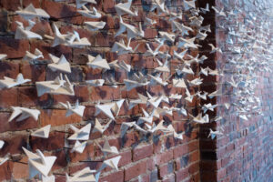 The title “Kiss the Joy as it Flies”, from William Blake’s poetry, aptly reminds us to notice the string of fleeting moments in life within the ultimate nature of impermanence.  This flock of 500 geometric abstracted birds plays with the balance within us of light and shadow, freedom and rigidity, movement and stillness. 

Photo credit: Cathie Ferguson