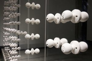 Two of the works in this series, Wall, and Gate seek to locate the body in a place of yearning anticipation, of wanting to reach for the other side, beyond the barrier and border. In the first of these, Wall, the room for the installation is divided into two halves. The viewer can enter into one side or the other, but once there cannot cross to the opposite side. The wall in this instance is composed of thirteen transparent panels with each one grounded in concrete building blocks, as if the foundation of home. The panels are scaled to the size of a door, and carry an array of white porcelain doorknobs suggesting an array of choice, yet none can turn, nor open passage to the other side. On one side of the installation a single white porcelain key is suspended from a golden thread, swaying and turning gently, yet just out of reach.
