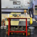 A counter-cum-workbench, tools and utensils act as both still life and kitchen in this installation. The smell of freshly cut pineapple seduces a viewer to slice, dice and pick to engage.

When is the last time you considered a pineapple? When is the last time you purchased a pineapple? Was it pre-skinned and diced? Prickly pineapples are slow laborious to cultivate. Each must be picked by hand, it is a cumbersome fruit. A worker wears full body armour when harvesting due to the knife-like spikes protruding from the tops of the fruit. A once-coveted Florida crop in the early 1900’s, the Dole company squashed the Florida-based industry by establishing its present day sites in Hawaii. The tropical fruit was a delicacy and the symbol for hospitality. At the end of a social gathering the host would ceremoniously cut the top off of the fruit, a sign that the party had ended. Still today, you can simply plant the top of a pineapple and cultivate your own. 

Abundance is key to this installation. The viewer is free to take, to participate.