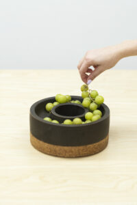 Photo Credit: Reba Jenson
The horderves and chaurceturie set are 8.25” wide and 4” tall, made of ceramic and cork; the piece aims to be functional in either orientation. The ceramic component is a double-walled thrown form that dips in the middle to create a rounded interior bowl to hold Horderves. When upright, the piece can be dual as a cracker and dip tray. The cork base continues to match the profile of the ceramic components. While the high-density cork performs better than hardwood, allowing it never to warp, it also provides a contrasting color pallet to the matte black. The collection celebrates sharing moments with others while focusing on how to encourage comfort and convenience for routine needs.