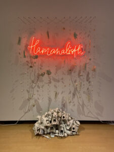 View with lights on of Tlamanalistli (sacrifice) showing details. Porcelain, neon, plexi: thread red neon text, loose porcelain 'curtain' of pills, flowers, and masks, flowing into pyramid of porcelain dipped skulls and thistles