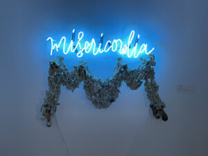 neon, porcelain, plexi, resin: blue neon text in Italian above a garland-style sculpture of porcelain pieces on plexi. Porcelain is soda-fired slip-dipped thistles, lilacs, stargazer lilies, and ballet pointe shoes, all in various shades from white to blue to grey, adhered with resin to plexi. 
Miseria et Misericordia (misery and mercy), pain and beauty, are two concepts that exist in constant companionship for women. The title is a reference to a speech by Pope Francis.
