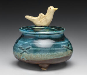 This wheel thrown jewelry jar has a hand pinched bird attached to top for a handle. Jar is decorated and altered with pattern on side by paddling. The feet are handbuilt and pinched instead of trimmed. It is glazed with hand mixed glazes and fired to cone 6 in oxidation with a 20 minute hold.