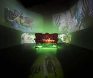 2021, 6 Projected Video, Couch, Terracotta. 

While the north space focuses on the Diva and the performance of gender and identity, it dehumanizes me, the south gallery works to re-humanizes me. The variation of close ups, angles, and subject matter works together to show a raw side of humanity: human desires, human actions, and sexuality. The south space exists within the past and as a transition out of the present. There is an undeniable presence of a stasis and waiting by the lounging bodies, and the never ending tapping of stiletto nails. While this space lies mostly within the realm of the interior, the acknowledgement of the viewer through the audio as well as the moments of locking eyes with the camera searching for eye contact on the other side speaks to a desire to be seen, foreshadowing what is to come within the near future.